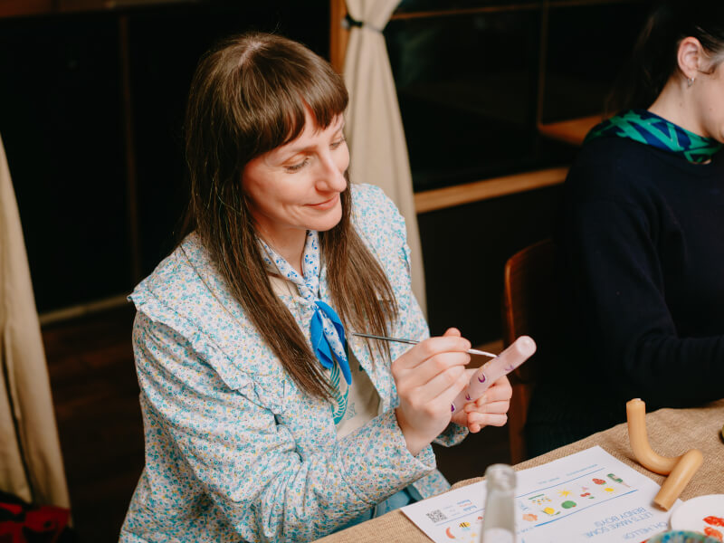 A woman is sitting at a table painting small motifs on a pink candle